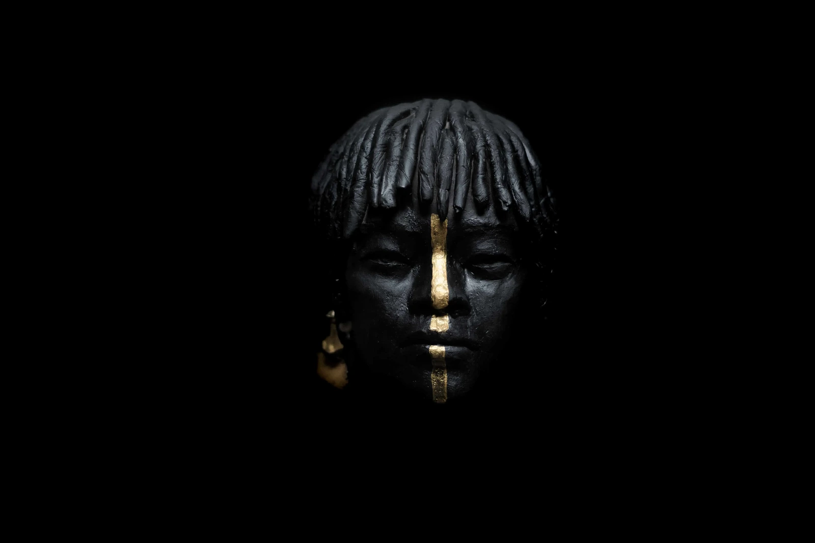 Sculpture de Terres Ethniques photographiée sur fond noir, visage marqué d'une ligne de feuille d'or